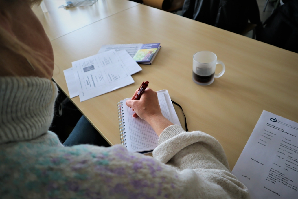Zum Vortrag Notizen machen Eine Teilnehmerin macht sich in einem kleinen Ringbuch Notizen. Auf dem Tisch liegen verschiedene Unterlagen. Auch ein Tasse Kaffee steht dabei.