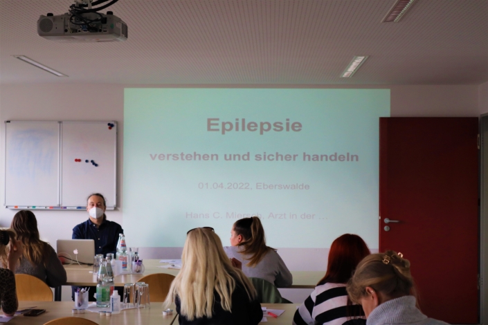 Das Schulungsthema – Epilepsie Auf der großen Leinwand ist das Schulungsthema Epilepsie verstehen und sicher handeln. Der Referent hat eine Maske auf. Vor ihm sitzen fünf Teilnehmerinnen.