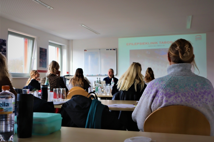 Blick von hinten über die Schulungsteilnehmer:innen Ein Blick von hinten auf den Referenten, der vor einer großen Leinwand mit einer PowerPoint-Präsentation sitzt. Davor sind die Rücken mehrerer Teilnehmerinnen zu sehen.