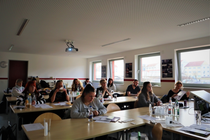 So sieht der Referent die Schulungsteilnehmer:innen Blick von vorn in den Schulungsraum. Insgesamt sind vier Tischreihen mit insgesamt 12 Personen zu sehen. Jeder zweite Stuhl ist besetzt. Rechts sind vier Fenster.