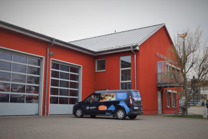 Das Fahrzeug der Lebenshilfe steht vor dem Feuerwehrgebäude Vor den beiden Toreinfahrten des roten Feuerwehrgebäudes steht das Essen-Lieferfahrzeug der Lebenshilfe Barnim. Der Giebel des Feuerwehrgebäudes ist ebenfalls zu sehen.