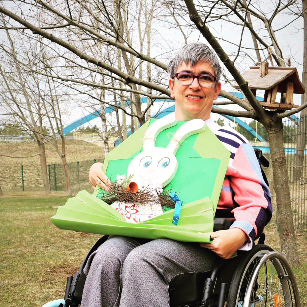 Beatrice mit unserer handgemachten Osterdekoration Beatrice steht mit ihrem Rollstuhl auf der Wiese vor unserem Vogelhaus. Auf dem Schoß hat sie die handgemachte Osterdekoration: Ein weißer Holzosterhase sitzt in einem großen, stilisierten hellgrünen Briefumschlag. Am Hals trägt er ein braunes Glöckchen.
