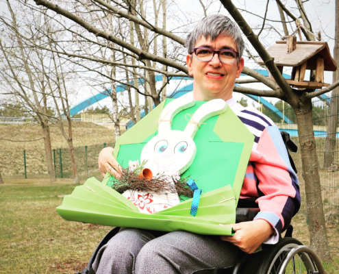 Beatrice steht mit ihrem Rollstuhl auf der Wiese vor unserem Vogelhaus. Auf dem Schoß hat sie die handgemachte Osterdekoration: Ein weißer Holzosterhase sitzt in einem großen, stilisierten hellgrünen Briefumschlag. Am Hals trägt er ein braunes Glöckchen.