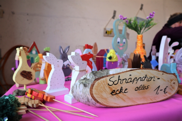Osterbasar in der Werkstatt – Schnäppchenecke Auf einem Tisch mit lila Tischdecke ist auf einem länglichen Stück Birkenholz der Aufdruck "Schnäppchenecke alle 1,- Euro" zu lesen. Dahinter stehen verschiedene bemalte Hasen, Kücken und Hühner.