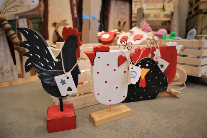 Osterbasar in der Werkstatt – der ‚Aufbau beginnt Vor mehreren Holzkisten, die mit österlichen Deko-Artikeln gefüllt sind steht ein weißes Holzhuhn mit roten Kamm und Schnabel. Es wird eingerahmt von zwei schwarzen Deko-Hähnen.
