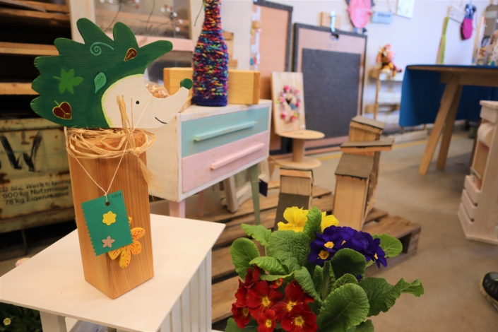 Osterbasar in der Werkstatt – auch Nistkästen gehören zum Angebot Auf einem weißen Holztischen steht ein grüner Igel. Dahinter ist eine bunte Kommode zu erkennen, auf der eine Flasche mit bunter Strickhülle steht. Davor befinden sich auf einer Palette mehre Nistkästen aus Holz. Rote, blaue und gelbe Primeln im Vordergrund bilden einen farbigen Tupfer.