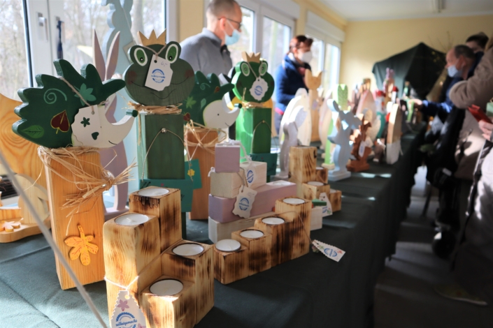 Sonnenhof Osterbasar – auch Igel und Kerzen gehören dazu Im Vordergrund stehen Holz-Kerzenhalter in verschiedenen Größen mit weißen Teelichtern. Dahinter sind Holzfiguren in Form von Igeln und Fröschen aufgestellt. Im Hintergrund sind zwei Mitarbeiter:Innen der Werkstatt und einige Kauf-Interessierte zu sehen.