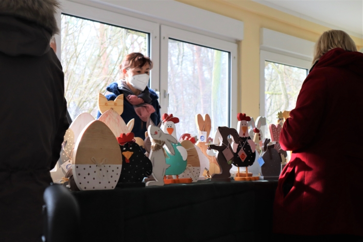 Sonnenhof Osterbasar – bunte Hühnerschar mit Osterhasen Auf einem langen Tisch stehen verschiedene Hühner, Hasen und Ostereier aus Holz. zum Verkauf. Hinter dem Tisch steht ein Mitarbeiterin der Werkstatt, davor einige Kundinnen.