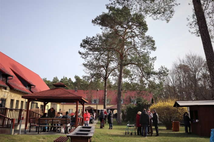 Osterbasar im Sonnenhof – Zeit zum Verweilen im Außengelände Das Außengelände vom Sonnenhof. Auf einer großzügigen Rasenfläche mit mehreren großen Kiefern stehen ein Holzpavillon und ein Holzhäuschen. Auf dem Gelände stehen, gehen und sitzen viele Menschen, einige mit Rollator, andere mit Rollstuhl. Am Holzhäuschen kann man Kaffee trinken. Das Außengelände wird teilweise von den Gebäuden des Sonnenhofes begrenzt.
