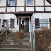 Eine junge Frau fegt vor dem Giebel eines 'Fachwerkhauses Gestrüpp eine Betontreppe herunter. Diese hat links und rechts ein graues Geländer.
