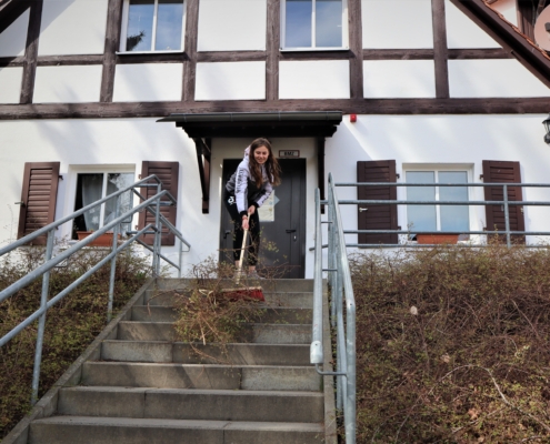 Eine junge Frau fegt vor dem Giebel eines 'Fachwerkhauses Gestrüpp eine Betontreppe herunter. Diese hat links und rechts ein graues Geländer.