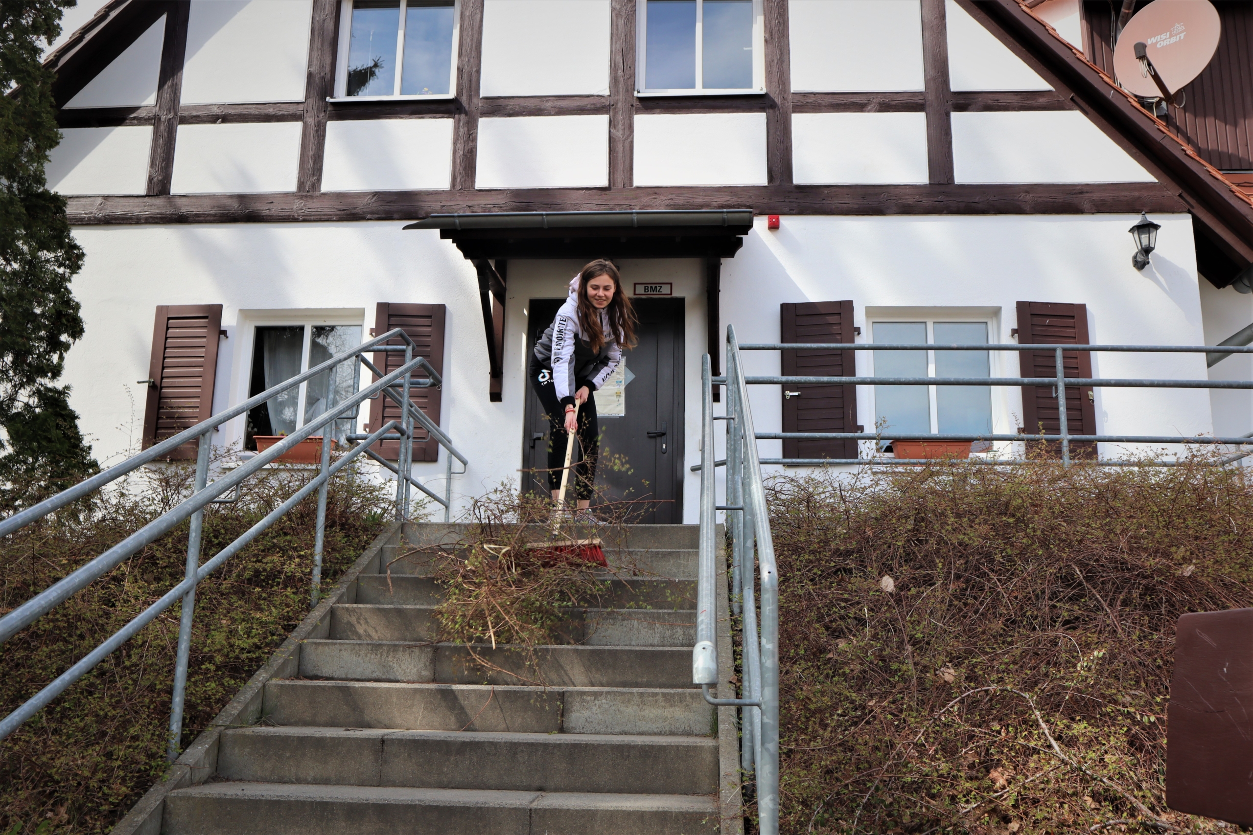 Eine junge Frau fegt vor dem Giebel eines 'Fachwerkhauses Gestrüpp eine Betontreppe herunter. Diese hat links und rechts ein graues Geländer.