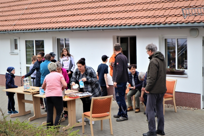 Vor dem flachen Verwaltungsgebäude der Wohnstätte "Heidehaus" versammeln sich etwa 15 kleine und große Teilnehmer:Innen am Frühjahrsputz zur wohlverdienten Pause. Auf zwei Tischen werden Kaffee und Kuchen aufgebaut.