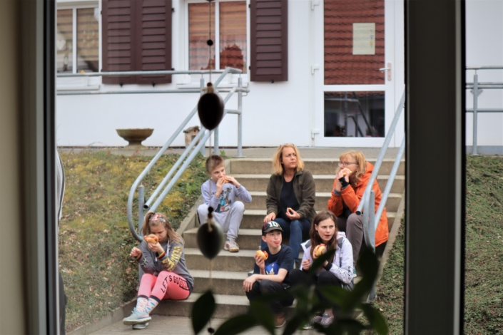 Auf der Treppe zu einem Wohnhaus haben es sich zwei Mädchen, zwei Jungen und zwei erwachsene Frauen gemütlich gemacht. Sie stärken sich mit Bratwurst im Brötchen.
