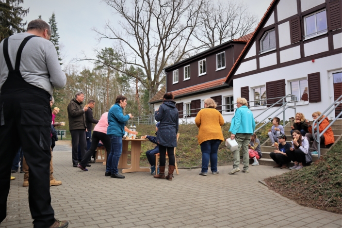 Noch ein Blick auf die versammelten Helferinnen und Helfer, Sie stehen und sitzen in weitem Rund auf grauem Betonpflaster um die beiden Tische und lassen sich Kaffee, Kuchen, Saft, Wasser und Herzhaftes schmecken.