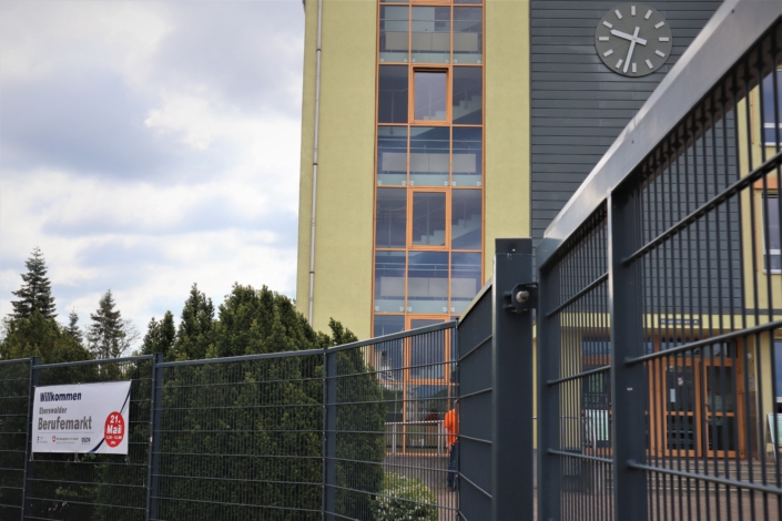 Am grauen Metallzaun des OSZ ist ein weißes Transparent mit der Aufschrift "Willkommen Eberswalder Berufemarkt" angebracht. Dahinter sind Teile der gelben Fassade und eine Uhr über dem Eingang zu sehen.