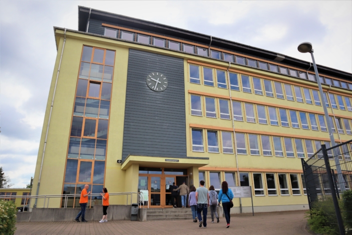Mehrere Besucher sind auf dem Weg zum Eingang des OSZ mit drei Stufen. Das moderne Gebäude mit vielen Fenstern hat vier Etagen und ein Dachgeschoss. Die Fassade ist ge.lb. Über dem, Eingang ist sie grau gestaltet. Im oberen Drittel befindet sich ein Uhr, die zwei Minuten nach halb neun anzeigt. Links führt eine Rampe zum Eingang. Davor stehen zwei Personen, die rauchen..