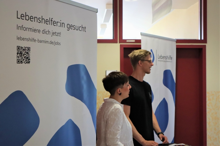 Unser junges Beraterpaar steht vor den beiden Rollups mit dem Logos der Lebenshilfe und dem Slogan "Lebenshelfer:in gesuicht".