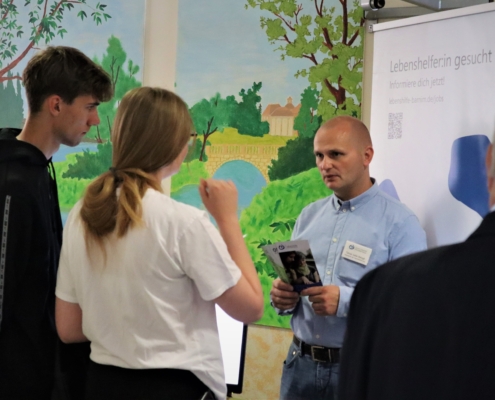 Unser Personalleiter, Herr Sallai-Balogh, hält einen Flyer in seinen Händen, während er sich mit einem jungen Mann und einer Frau unterhält. Am rechten Bildrand sind noch Kopf und Rücken eines älteren Mannes zu sehen.