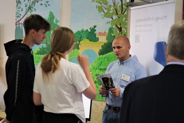 Unser Personalleiter, Herr Sallai-Balogh, hält einen Flyer in seinen Händen, während er sich mit einem jungen Mann und einer Frau unterhält. Am rechten Bildrand sind noch Kopf und Rücken eines älteren Mannes zu sehen.