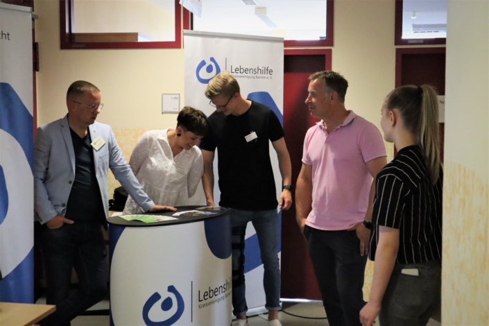 Eine Besucherin steht hinter dem Stand zwischen Herrn Lutter und dem jungen Mitarbeiter der Lebenshilfe. Davor stehen ein Mann und eine junge Frau mit blondem Pferdeschwanz.