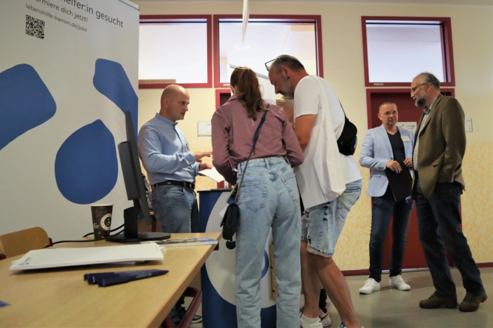 Herr Sallai-Balogh berät eine junge Frau und einen Mann. Im Hintergrund ist Herr Lutter, Geschäftsführer, mit Landrat Daniel Kurt im Gespräch.