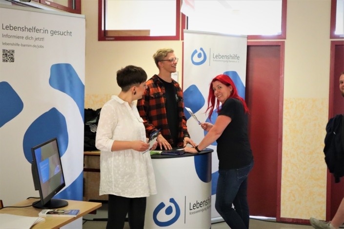 Am Stand der Lebenshilfe steht ein junge Frau mit langen roten Haaren, die in ihrer rechen Hand einen Flyer hält. Die beiden jungen Mitarbeiter:innen stehen hinter dem Stand.