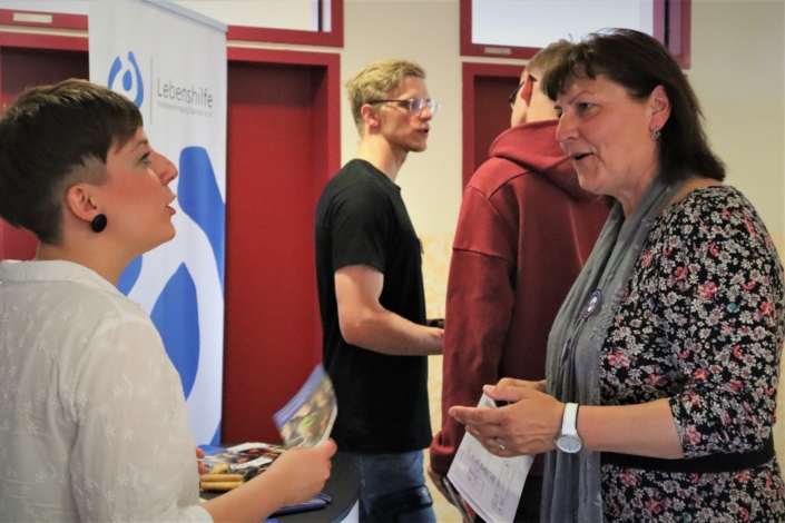 Im Vordergrund spricht die junge Mitarbeiterin der Lebenshilfe mit Frau Lüssow (Bereichsleiterin Ambulante Dienste der Lebenshilfe). Dahinter gibt der junge Mitarbeiter der Lebenshilfe einem jungen Mann Auskunft.