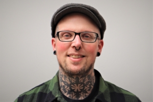 Kunstkurs - Björn Roters Porträt von Björn Roters, dem Leiter des Kunstkurses. Björn Roter hat eine graue Schirmmütze auf und trägt eine Brille. Er hat einen leichten Kinnbart, Tunnel-Ohrringe und ein grünes Hemd an. Der Hals ist tätowiert.