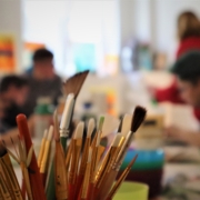 Den Vordergrund des Fotos bilden Pinsel in verschiedenen Größen. Der Rest des Raumes ist mit Abicht unscharf aufgenommen.