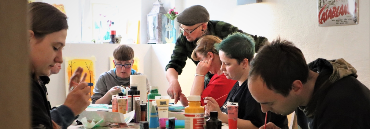Kunstkurs - alle an einem Tisch Auf dem Tisch stehen Farben, Pinsel und Wassergefäße. An der rechten Seite sitzen zwei Frauen und ein Mann. Auch an der unteren Tischseite sitz ein Kurteilnehmer, Björn Roters zeigt auf das Bild einer Frau am Ende des Tisches.