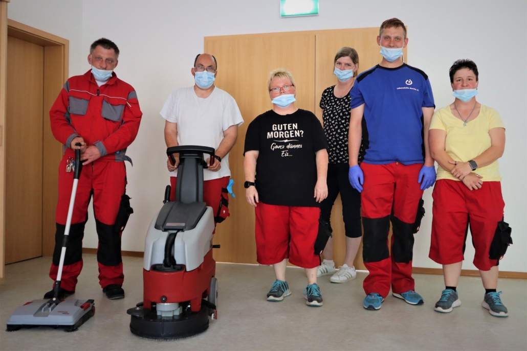 Unser Serviceteam - das sind wir Das Serviceteam hat sich mit insgesamt sechs Personen zum Gruppenfoto aufgestellt. Links stehen zwei Männer, die Reinigungsmaschinen in ihren Händen halten. Daneben stehen zwei Frauen, darunter die Gruppenleiterin. Den rechten Rand bildet eine Frau. Das Team trägt rote Arbeitshosen. und T-Shirts.