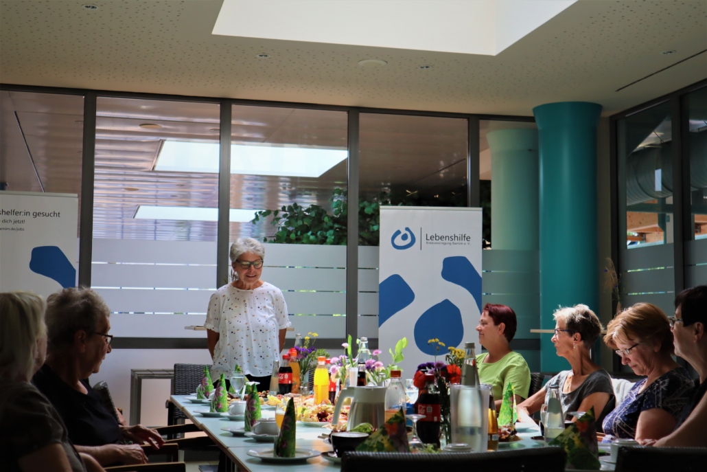 Dank an die Ehrenamtlerinnen und Ehrenamtler Im Bistro im Baff sitzen sechs Frauen an einem schön gedeckten Tisch. Die Vorsitzende der Lebenshilfe Barnim e. V. steht an einer Stirnseite und spricht zu den Anwesenden. Im Hintergrund ist ein Rollup mit dem Logo der Lebenshilfe zu sehen,