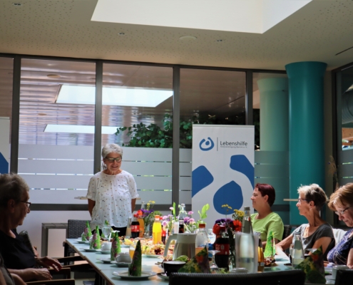 Im Bistro im Baff sitzen sechs Frauen an einem schön gedeckten Tisch. Die Vorsitzende der Lebenshilfe Barnim e. V. steht an einer Stirnseite und spricht zu den Anwesenden. Im Hintergrund ist ein Rollup mit dem Logo der Lebenshilfe zu sehen,