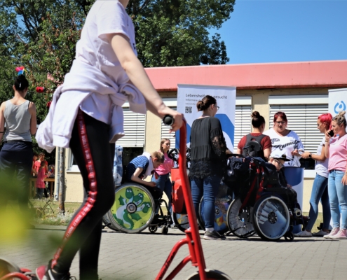 Eine Besucherin ist mit einem roten Roller auf dem Schulhof unterwegs. Dahinter stehen mehrere Kinder und Jugendliche im Rollstuhl am Stand der Schulbegleitung.