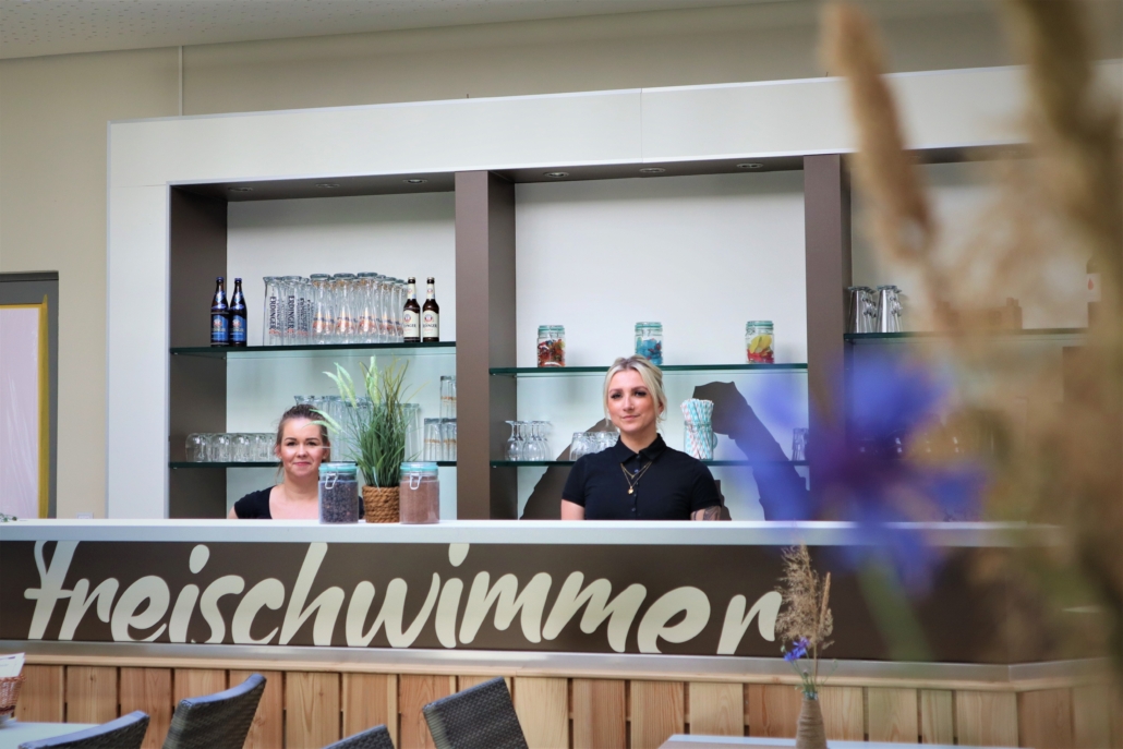 Sie sorgten für guten Service Die beidem freundlichen Restaurantfacharbeiterinnen stehen hinter dem Tresen, mit der Aufschrift Freischiwimmer. Das Wandregal hinter den Frauen ist mit verschiedenen Flaschen und Gläser bestückt.