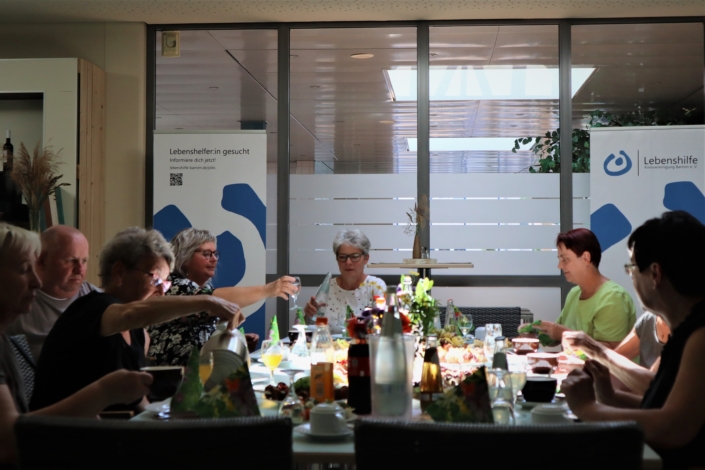 Gemütliche Runde Wieder ein Blick in die Runde am Tisch. An der Stirnseite sitzt Frau Winter. Hinter ihr stehen zwei Rollups mit den Logos der Lebenshilfe. Die anderen Teilnehmerinnen und Teilnehmer haben zum Teil Gläser in den Händen.
