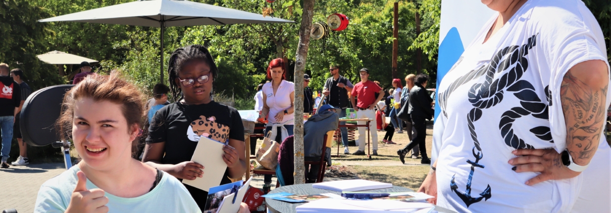 Seitlich fällt der Blick auf unseren Stand. Dahinter steht eine lachende Mitarbeiterin, davor eine junge Frau, die den erhobenen Daumen in die Kamera hält. Weitere Besucherinnen und Besucher halten sich im Hintergrund auf. Den Abschluss bilden grüne Büsch und Bäume.