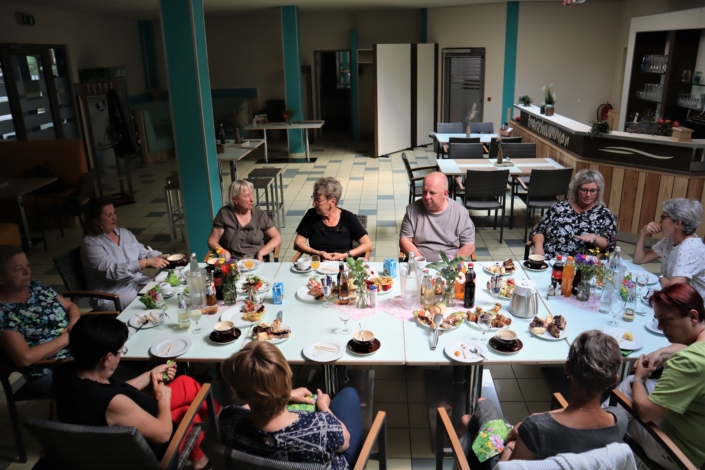 Alle an einer Tafel Ein Blick von schräg-oben auf die am Tisch sitzenden Ehrenamtlerinnen und Ehrenamtler. In der Runde sitzen zehn Frauen und ein Mann. Der Tisch ist schön gedeckt mit Kaffee, Kuchen, Getränken und einem kleinen Imbiss. Im Hintergrund ist der übrige Gastraum zu sehen.