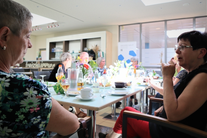 Dialog zwischen zwei engagierten Frauen Im Vordergrund tauschen zwei Frauen ihre Erfahrungen aus. Auch andere aus der Runde unterhalten sich im Hintergrund miteinander. Links vom Tisch ist ein Teil des Tresens sichtbar.