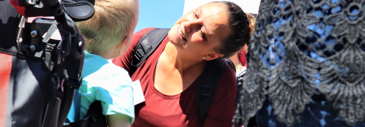 Eine Schulbegleiterin spricht mit einem blonden Jungen, der im Rollstuhl sitzt. Dabei geht sie in die Knie um dem Gesprächspartner in die Augen zu sehen.